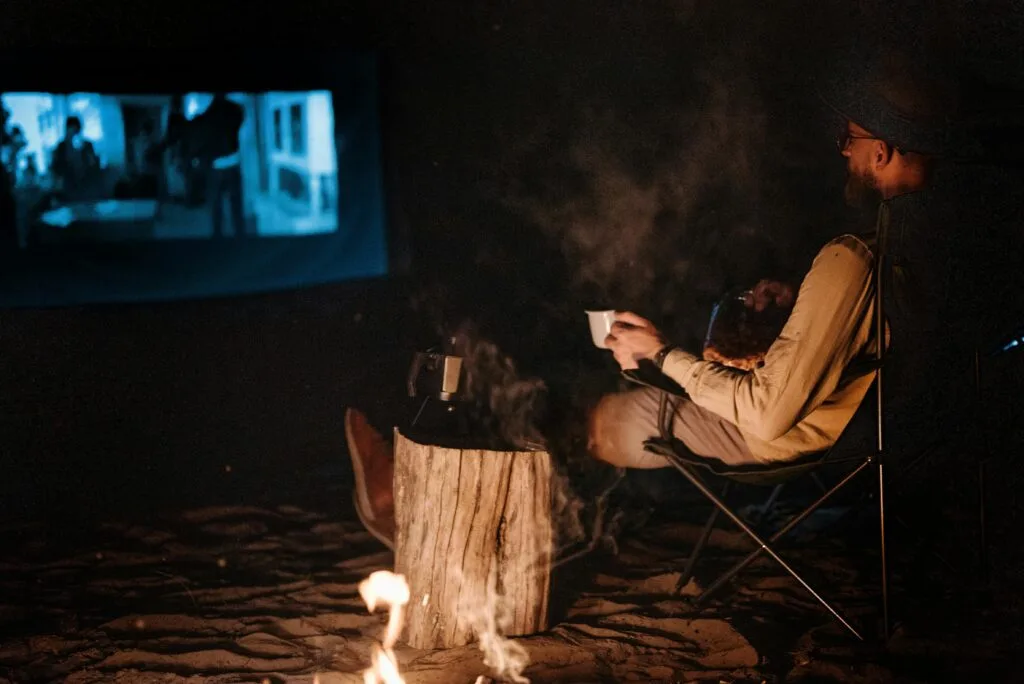 A man watching movie projected on the white curtain at the campsite